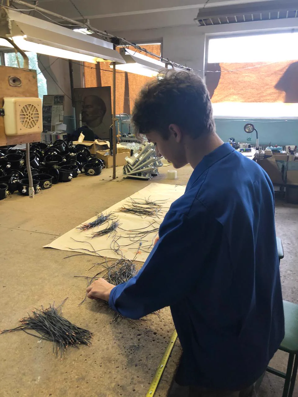 Me at a workshop table in a blue work coat, sorting bundles of wire, with rows of black lamp housings behind me.