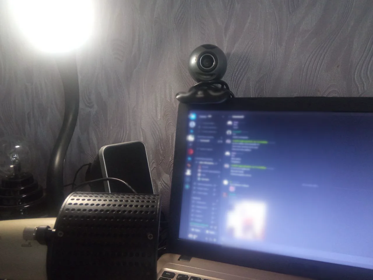 My first computer corner: a laptop with a blurred screen, a webcam, a speaker and a desk lamp against a grey wall.