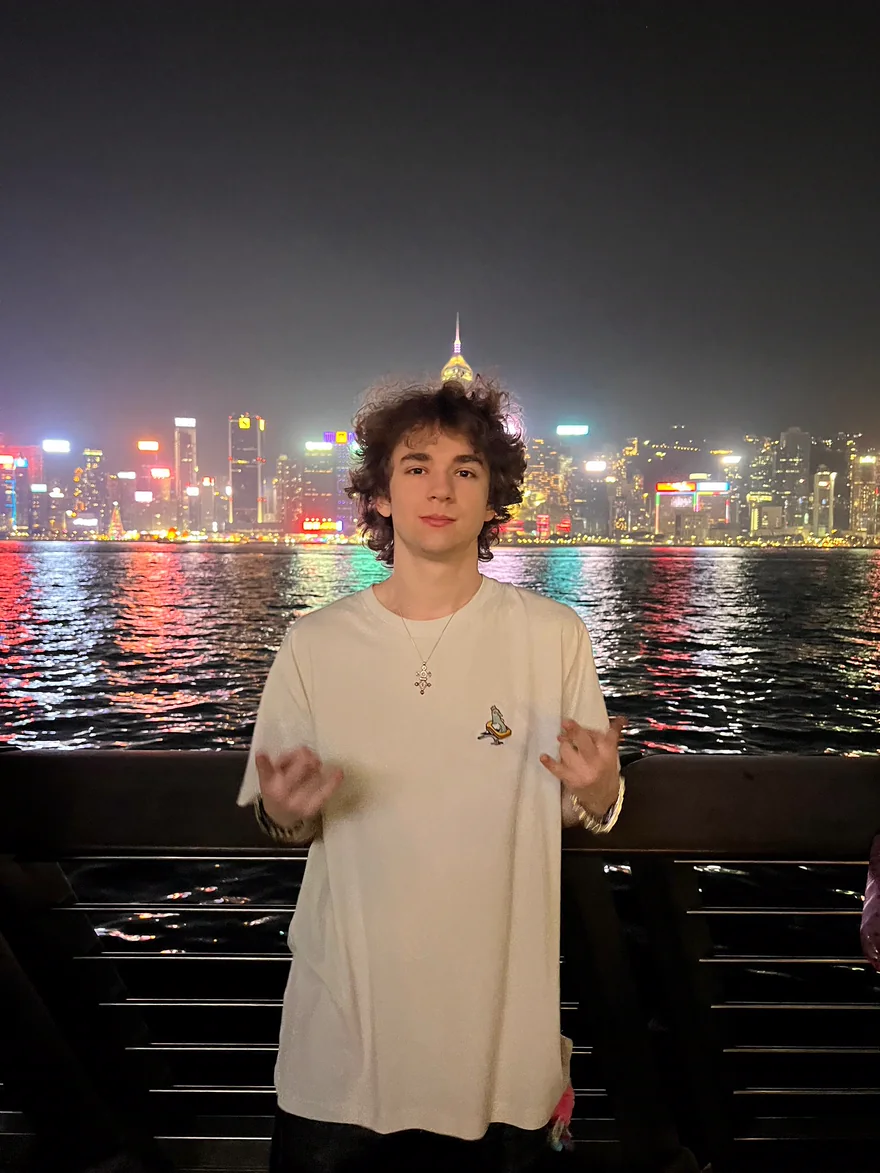 Me today, standing at night on a waterfront with the lights of a big city skyline across the water behind me.