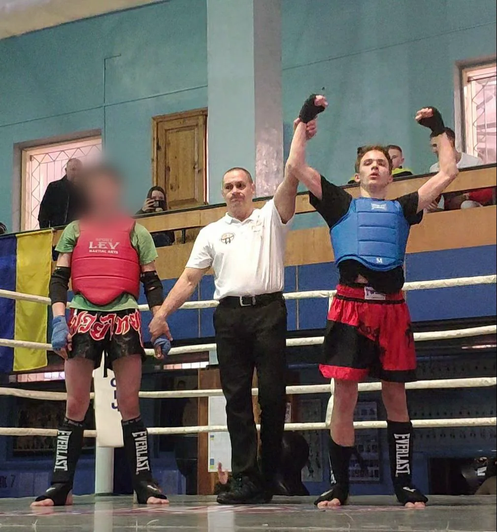 After a Muay Thai bout a referee raises the hand of a fighter in a blue chest guard and red shorts; his opponent stands on the left.