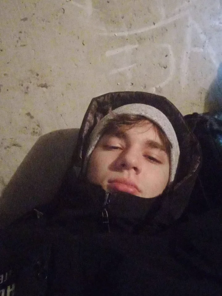 A teenager in a grey hat and a camouflage jacket holds a paper cup under a dark roof at night.