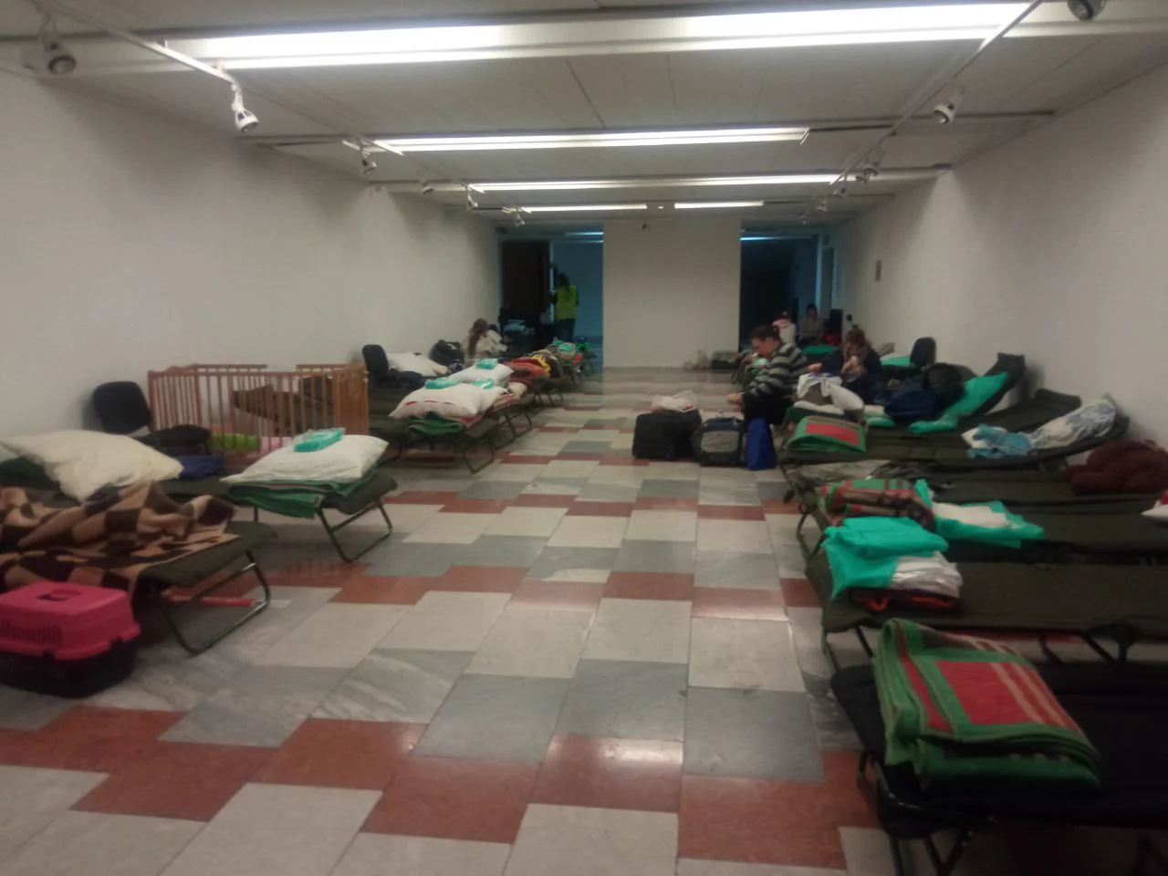 A long hall with rows of camp beds, blankets and bags; people sit or rest along the walls.