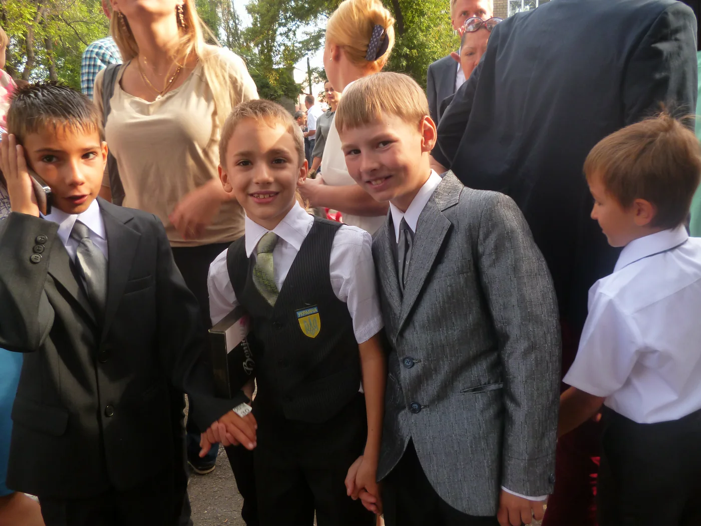 Boys in school suits and white shirts in a crowd outside, two of them smiling at the camera.