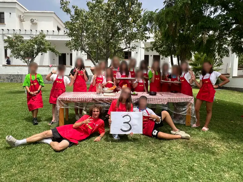A team in red aprons and red shirts behind a covered table on a lawn, in front of a white house; two of them lie in the grass holding a sign with a number three.