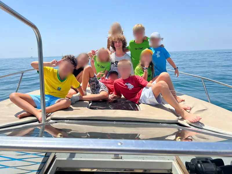 Children in bright T-shirts sit and lie together on the bow of a boat on a calm blue sea, some of them making peace signs, a helper with curly hair behind them.