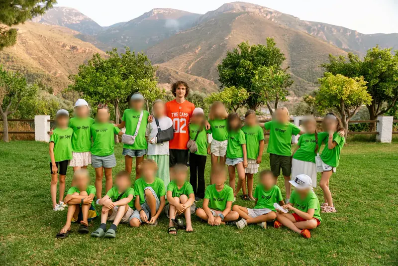 About twenty children in green camp T-shirts and one tall helper in an orange shirt stand and sit on the grass in front of mountains and orange trees.