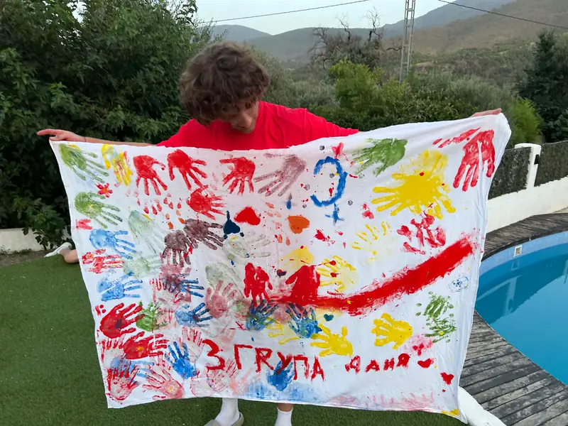 A teenager in a red shirt holds up a white sheet covered in coloured hand prints, with a group number and a name painted along the bottom, on a lawn next to a swimming pool.
