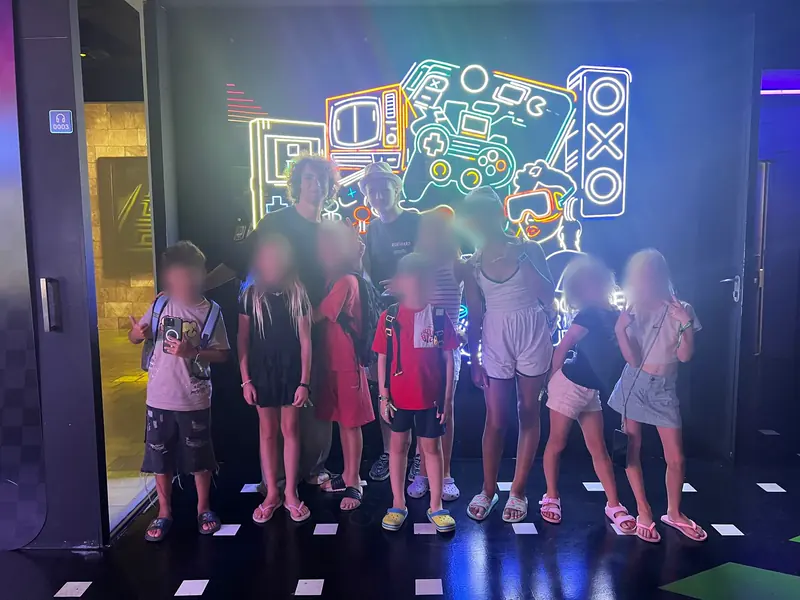 A group of children and two older helpers stand in a dark room in front of a neon drawing of game consoles.